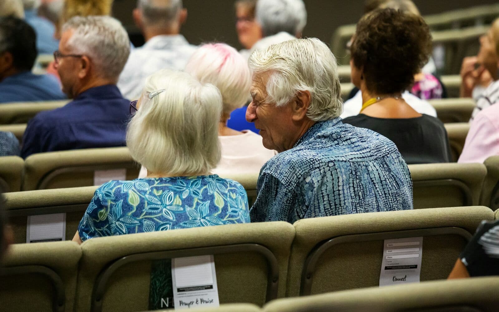 Des personnes âgées assises dans une salle, attentives, vues de dos.