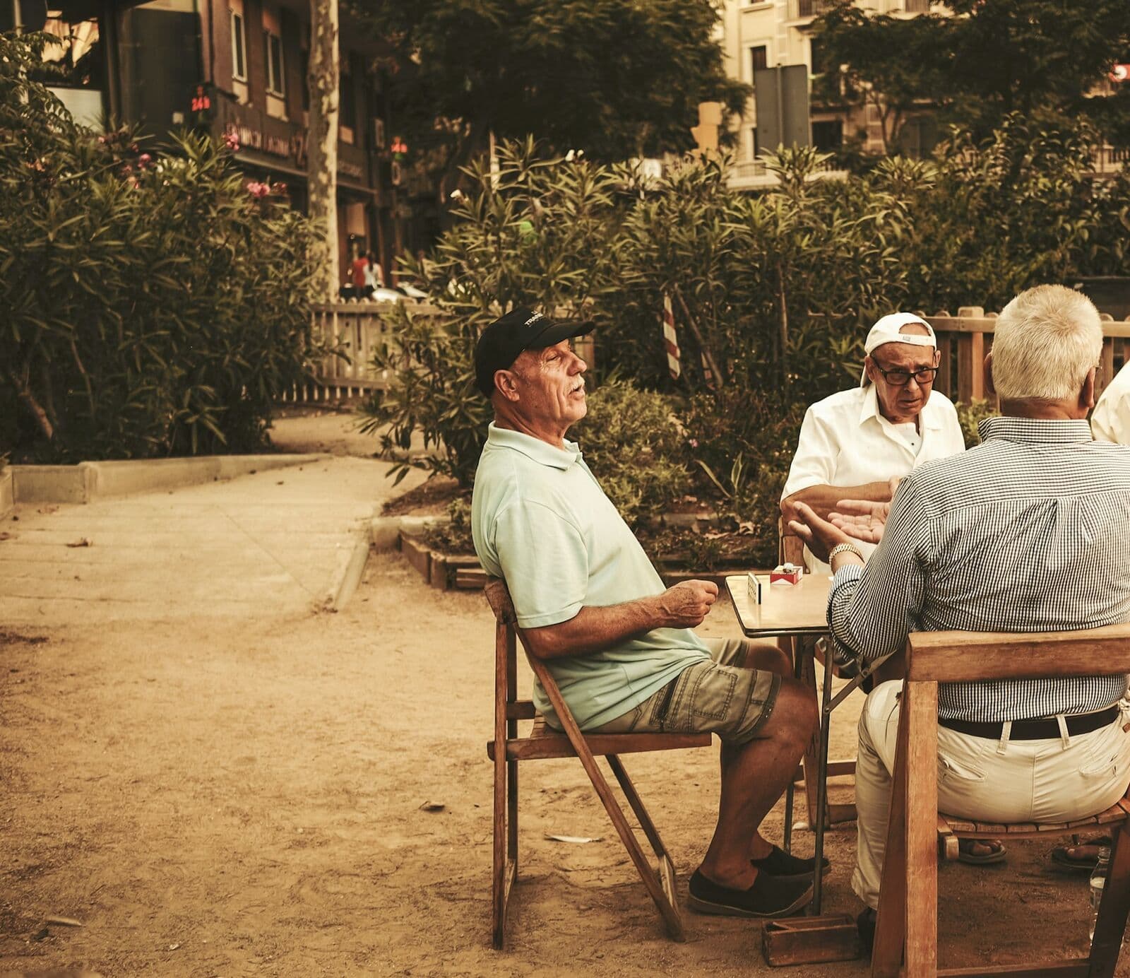 Des retraités discutent autour d'une table, en plein air, à l'ombre des arbres.
