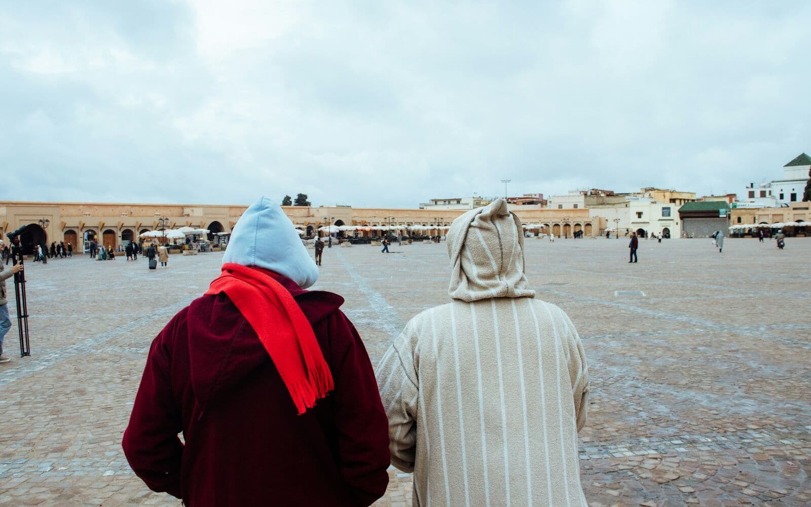 Deux personnes en djellaba marchent côte à côte sur une grande place au Maroc.